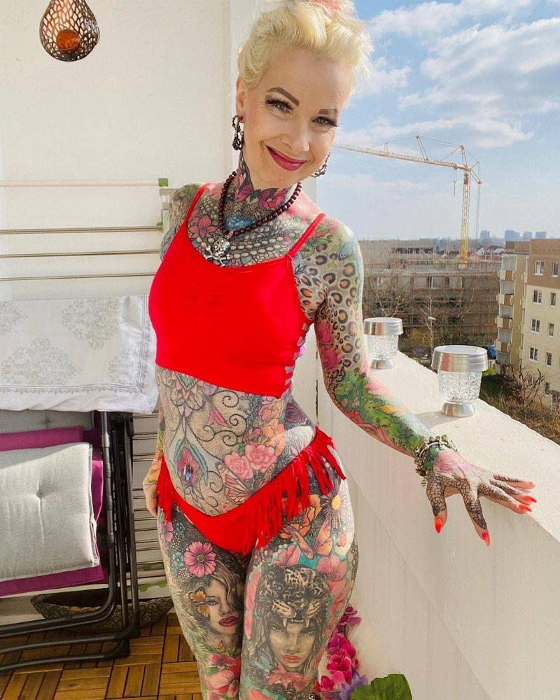 Tattooed woman stands in red two-piece swimwear on balcony, smiling
