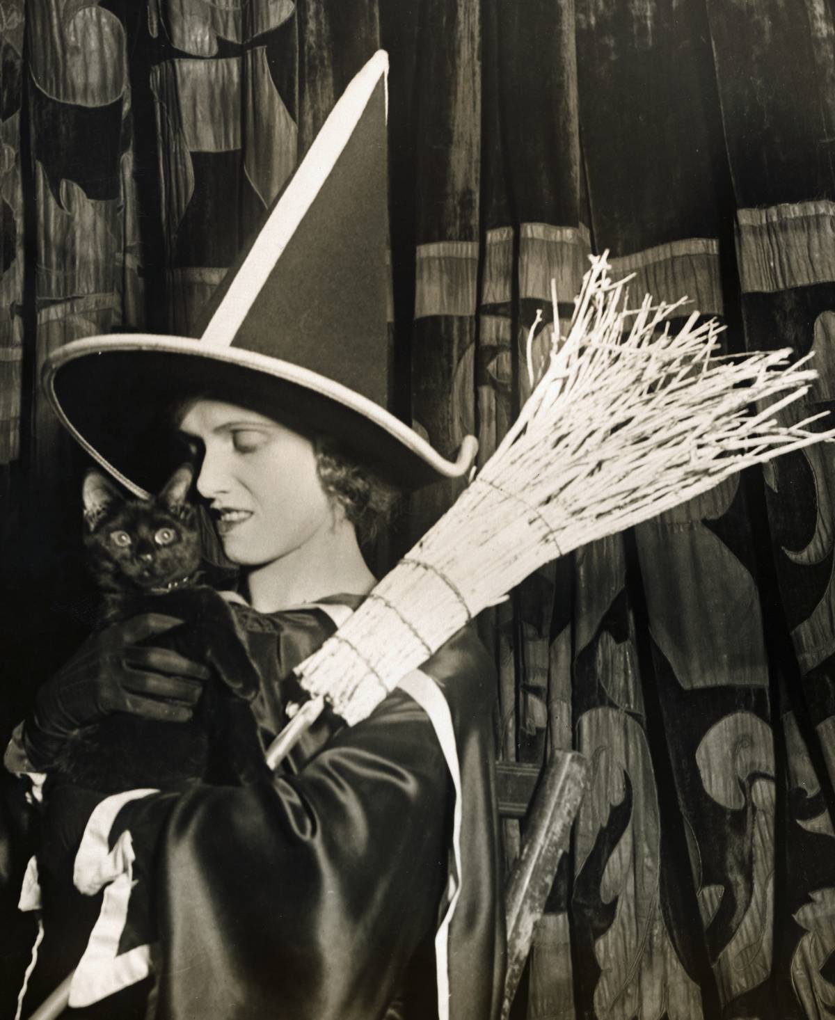 A woman wearing a witch costume, holding a straw broom and a black cat