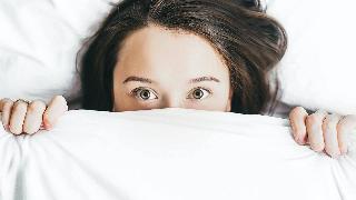 A girl lying in bed, holding the covers over her face with her eyes open.