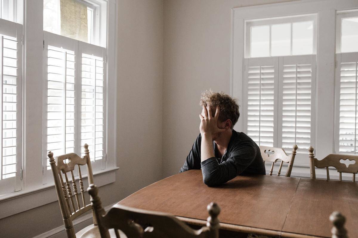 A man sitting alone at a kitchen table, his face resting in his hand.