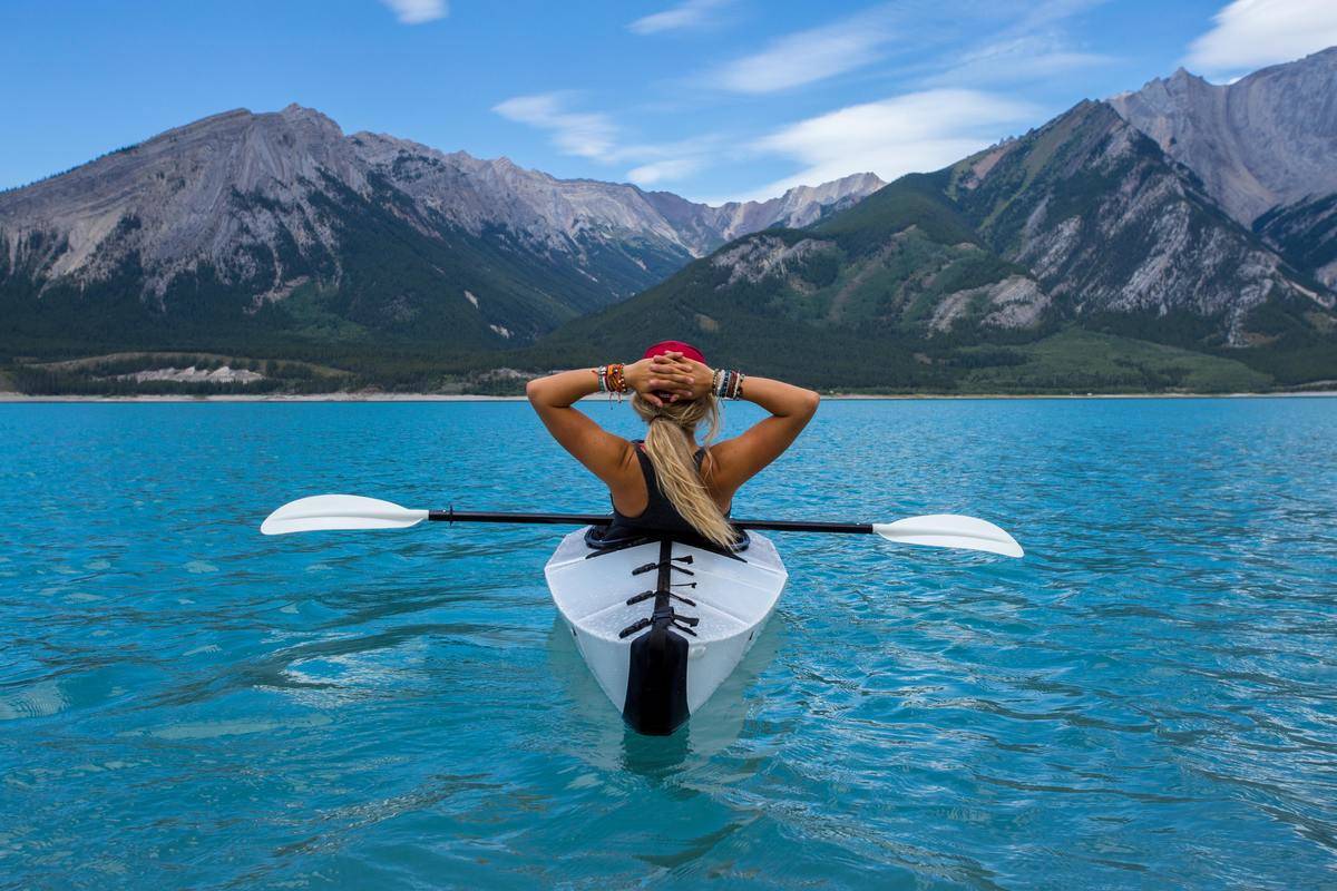 The back of a woman sitting in a canoe, floating in blue water with a mountain in the background.