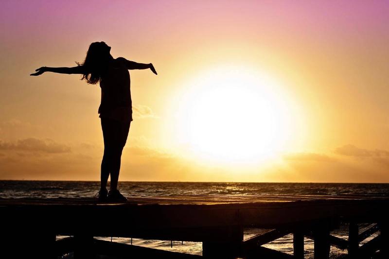 A woman standing near the ocean at sunset with her arms open wide and her head thrown back.