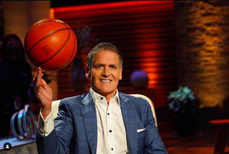 Mark Cuban balancing a basketball on the tip of his finger, smiling at the camera and wearing a blue suit.