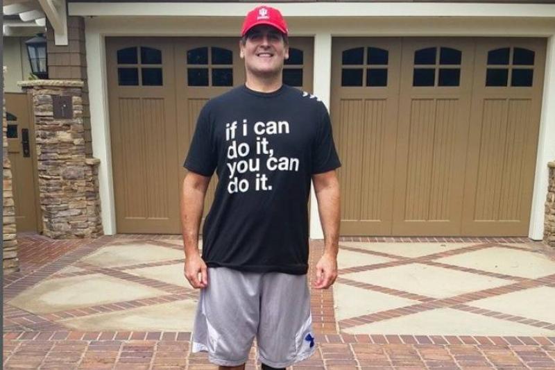 Mark Cuban standing on a driveway wearing a red hat and a black t-shirt that says, 