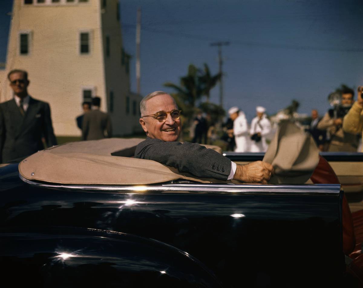 President Harry S. Truman riding in the back of a convertible with the roof off, smiling.