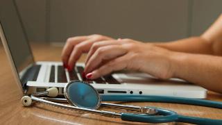 A woman typing on a laptop next to a stethoscope.