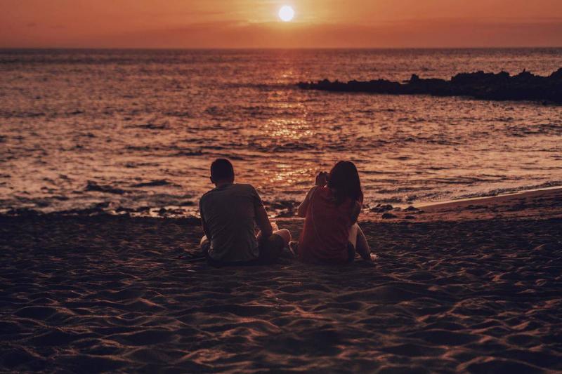 A young couple sitting on a beach at sunset.
