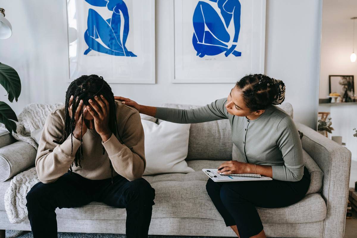 A man with his face buried in his hands, a woman comforting him while they sit on a white couch.