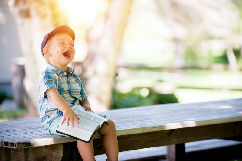 A little boy sitting on a wooden bench holding a book and laughing.