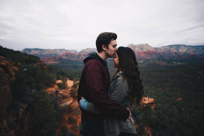 man kisses woman's forehead as they stand on a cliff