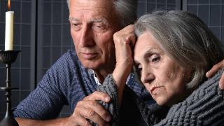 older man holds woman's arm as they sadly look at candle