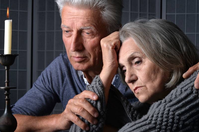older man holds woman's arm as they sadly look at candle