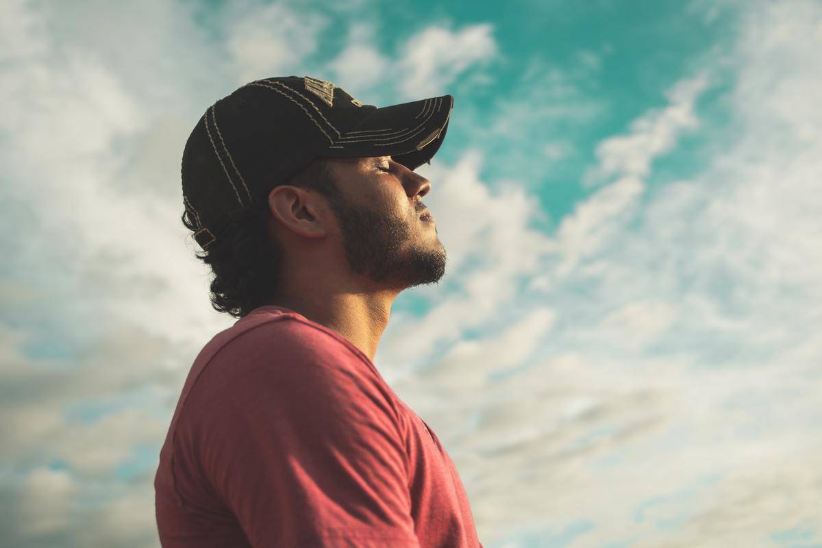 man-wearing-black-cap-with-eyes-closed-under-cloudy-sky