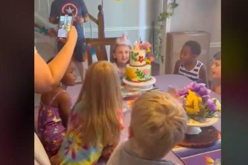 A group of kids sitting around a table singing happy birthday and looking at a cake.