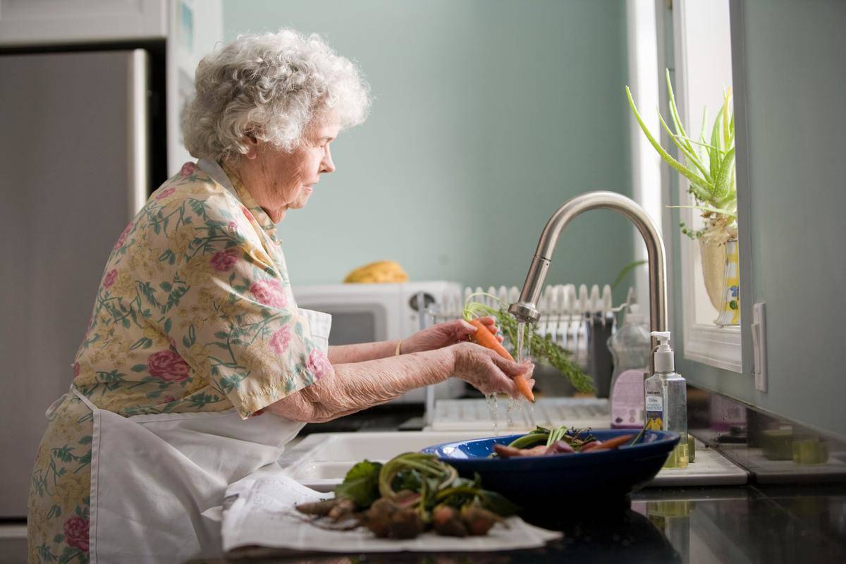 An elderly lady washing carrots at a kitchen sink.