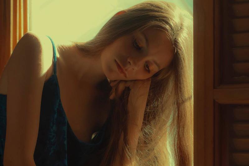 A woman with long blonde hair sitting at a sun-lit windowsill with her cheek resting on her hand.