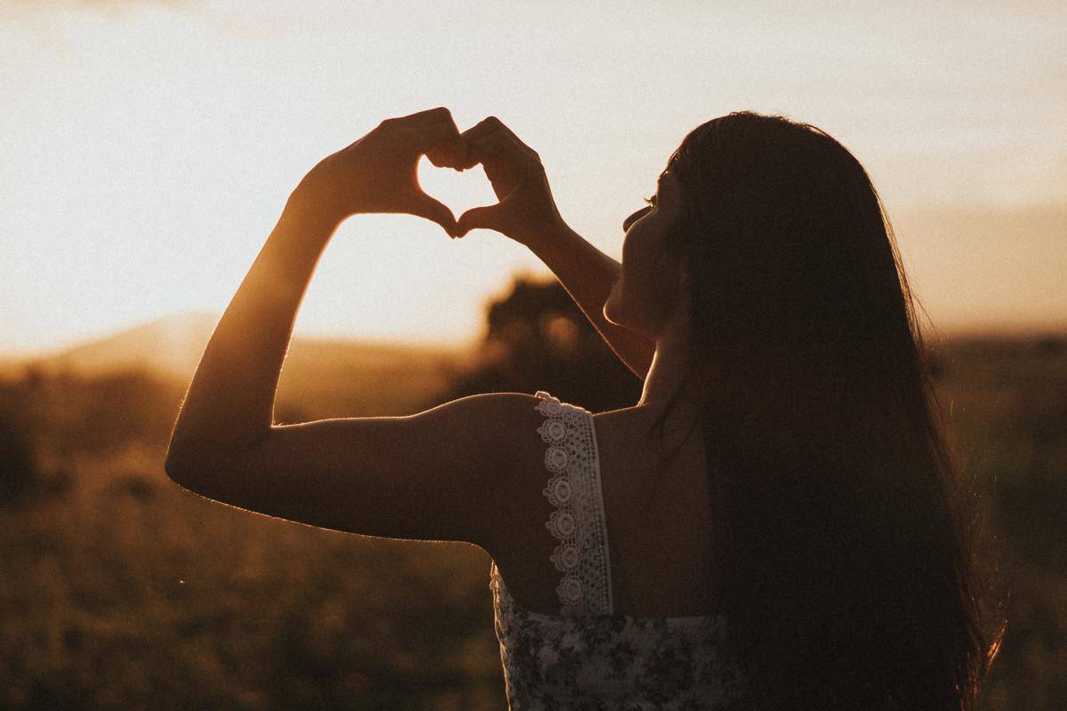 The back of a woman who is holding her hands up forming a heart, standing in front of a sunset.
