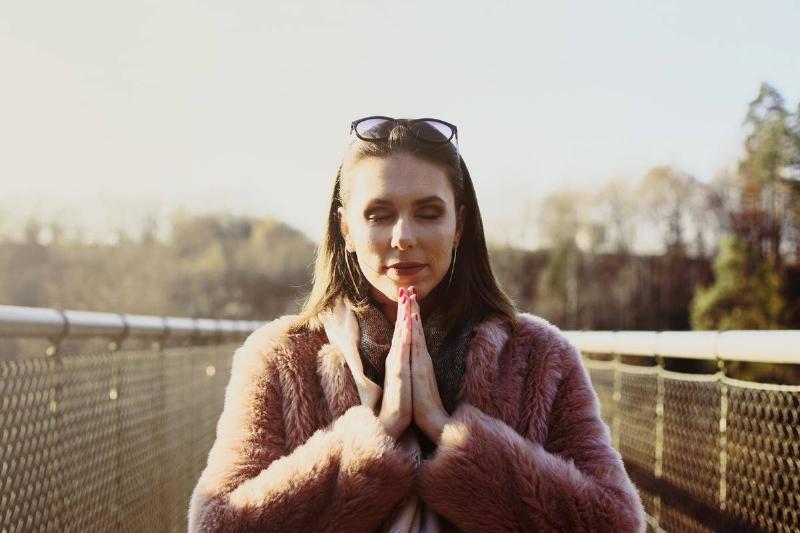 A woman wearing a pink furry jacket standing on a bridge, holding her hands together in prayer.