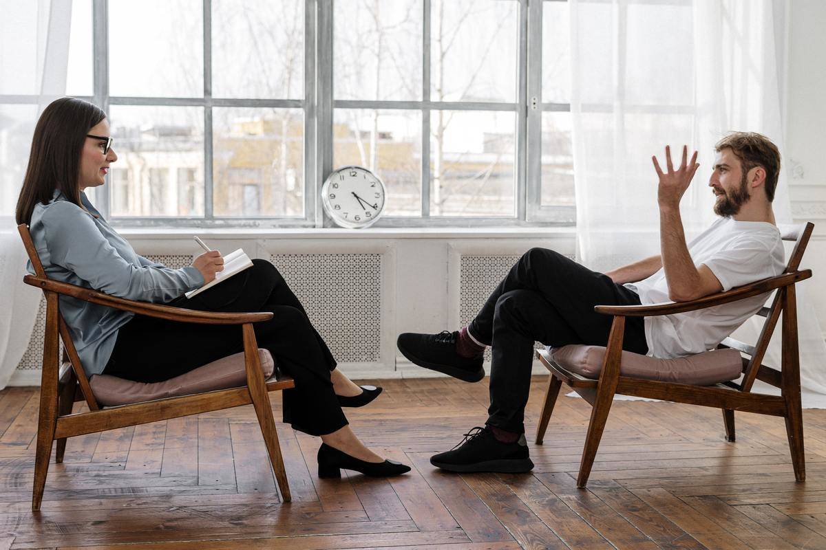 A man raises his hand in conversation at a therapy session while a woman takes notes.