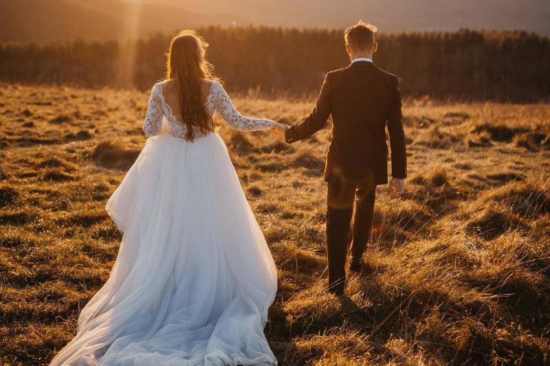 A man and woman walking hand in hand in a filed, wearing a wedding dress and suit.