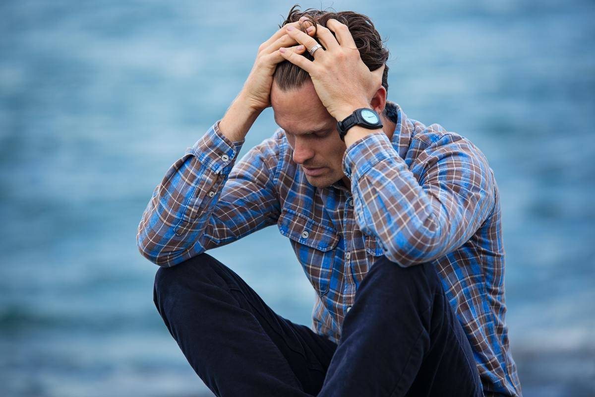 man sits by the water pushing his hair up looking concerned