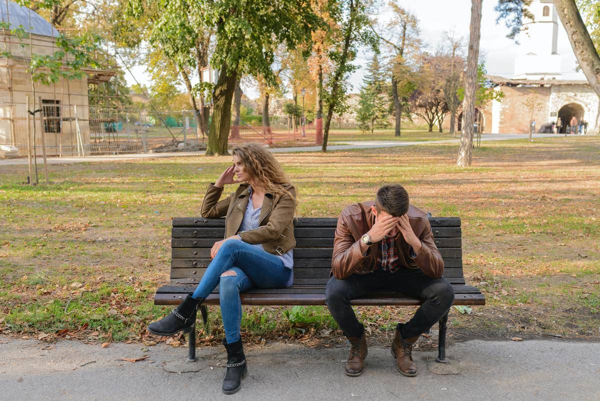 A man and woman sitting on a park bench, his head resting on his hands, her facing away from him in sadness. 