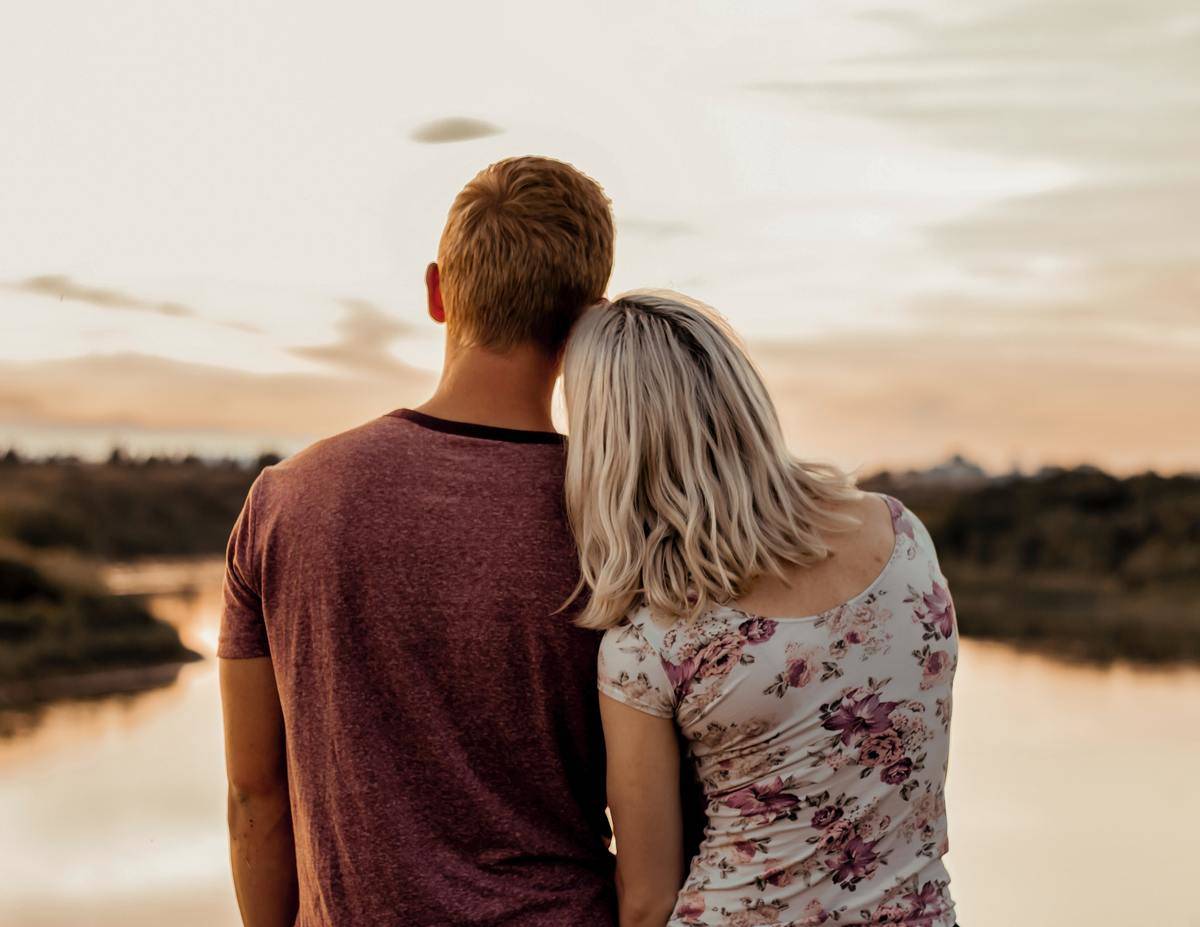 A young couple facing a lake at sunset, her head on his shoulder.