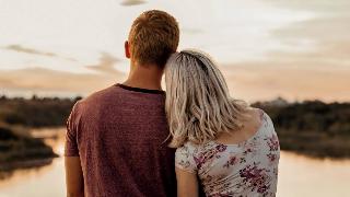 A young couple facing a lake at sunset, her head on his shoulder.