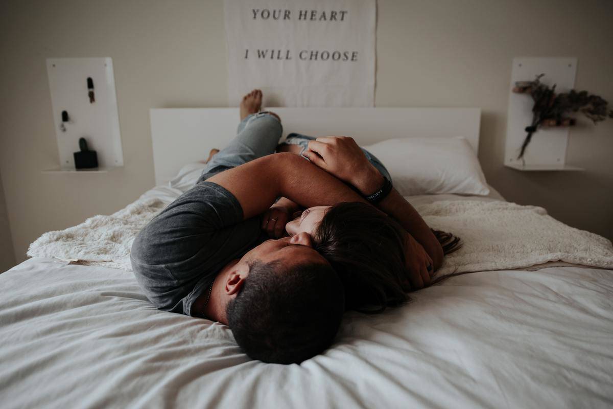 A young couple laying on a white bed cuddling, with a poster on the wall reading 