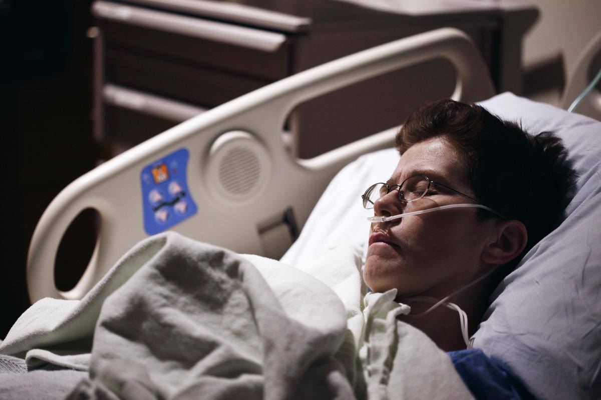 A person laying in a hospital bed, eyes closed and a tube in their nose.