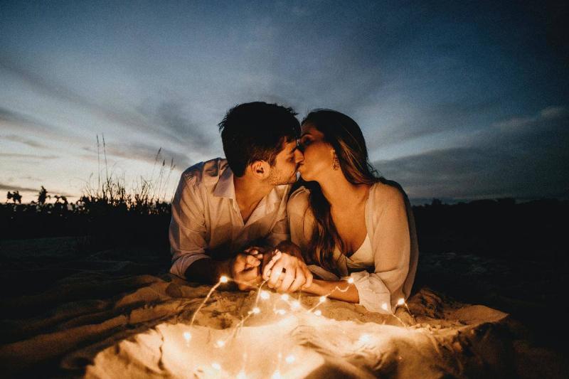 A man and woman sitting in front of string lights, kissing.