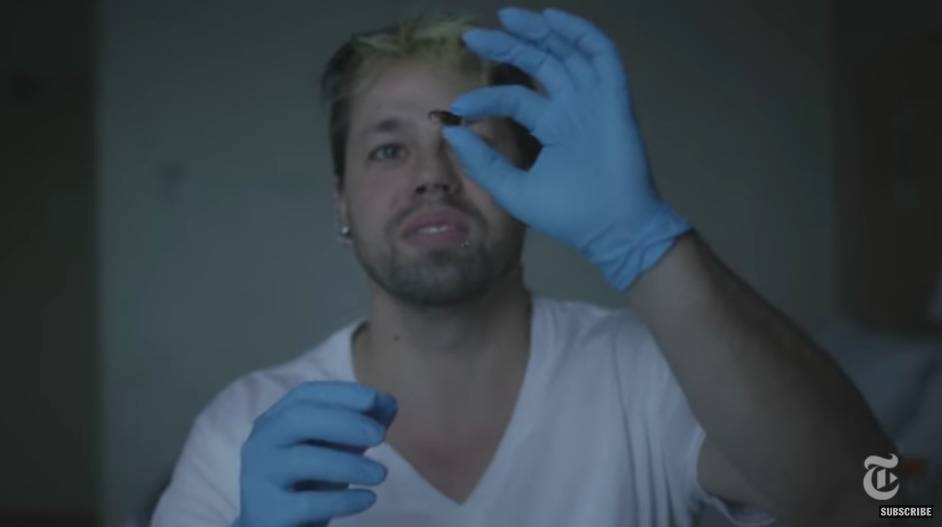 A man wearing blue latex gloves holding up a pill and examining it.