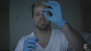 A man wearing blue latex gloves holding up a pill and examining it.