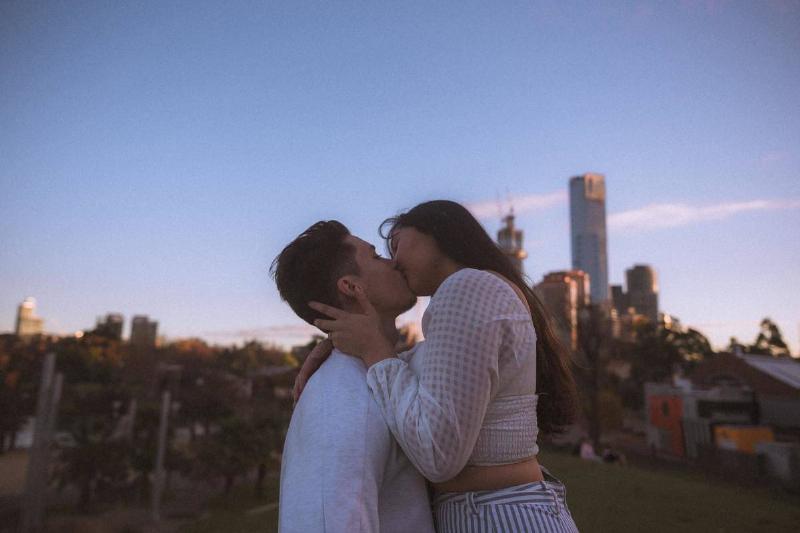 A guy and girl kissing outside in front of buildings.