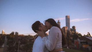 A guy and girl kissing outside in front of buildings.