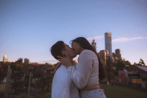 A guy and girl kissing outside in front of buildings.