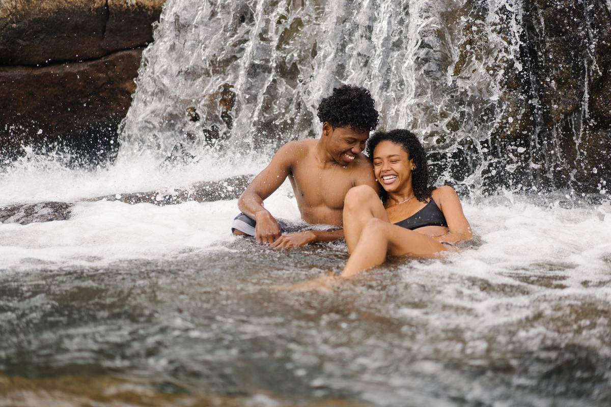 A young guy and girl laughing, sitting under a waterfall in bathing suits.