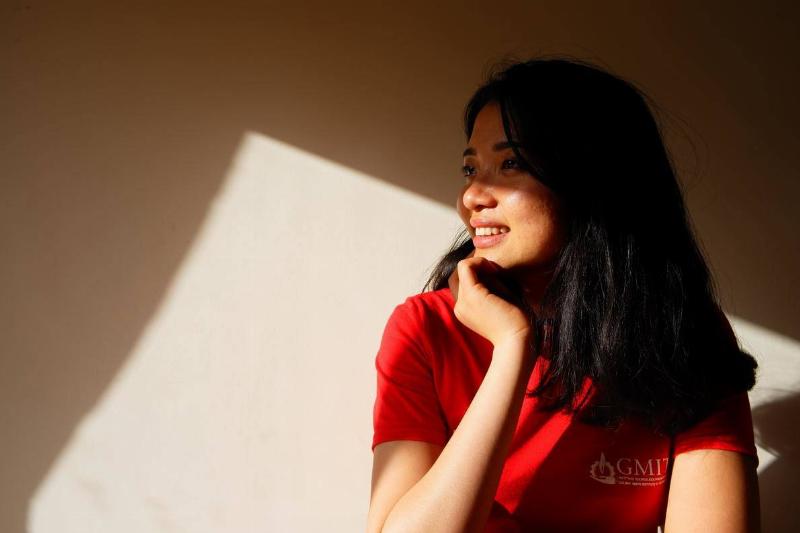 A woman in a red shirt smiling in front of a shadowed wall.