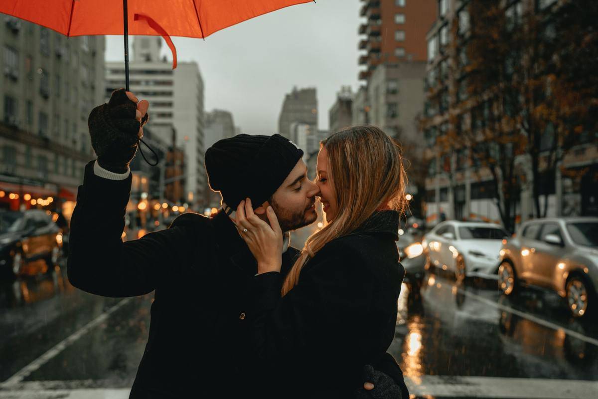 A young couple kissing underneath a red umbrella in front of a busy city street. 