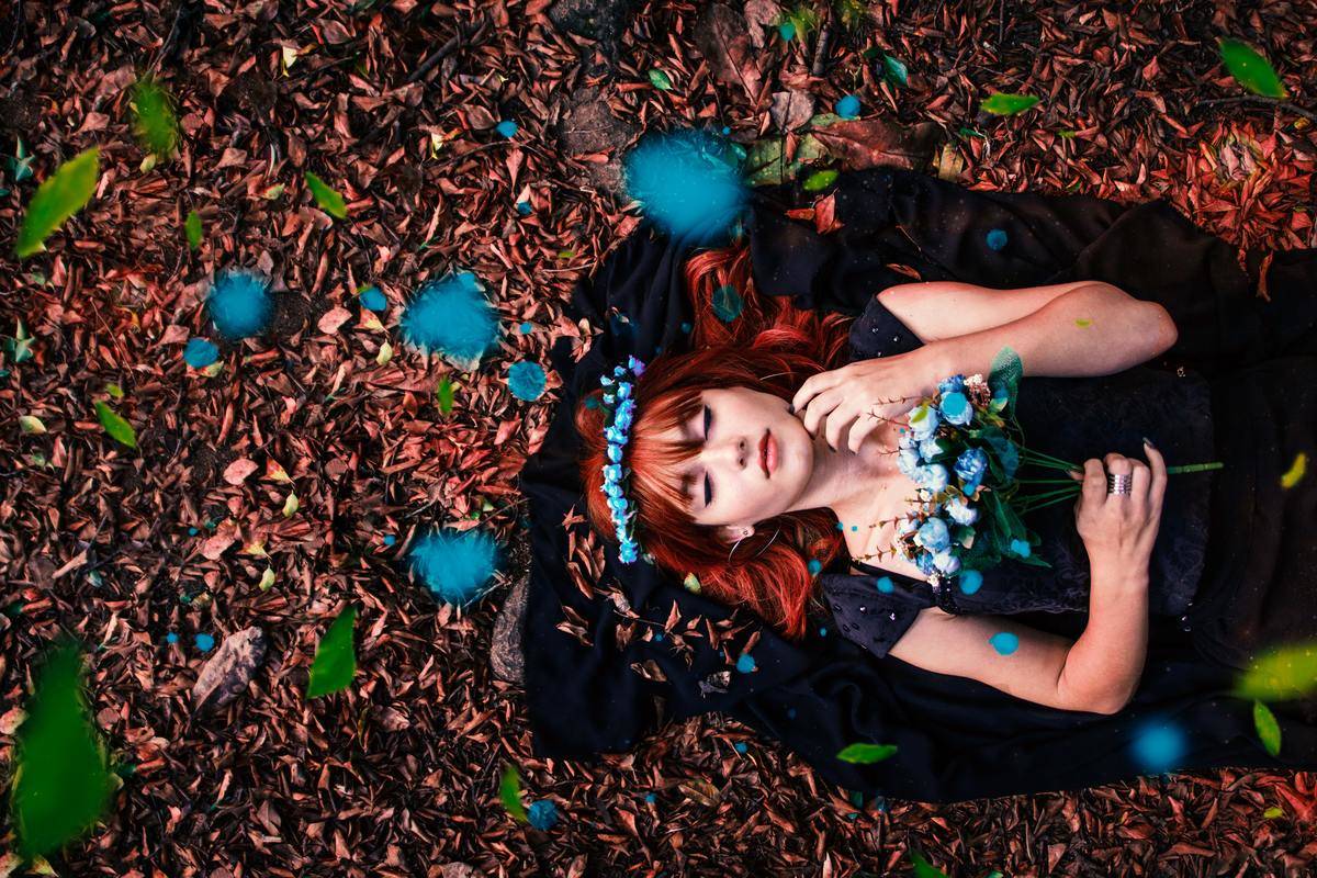 woman-in-black-dress-holding-bouquet-of-blue-flowers-while-lying-on-dried-leaves-