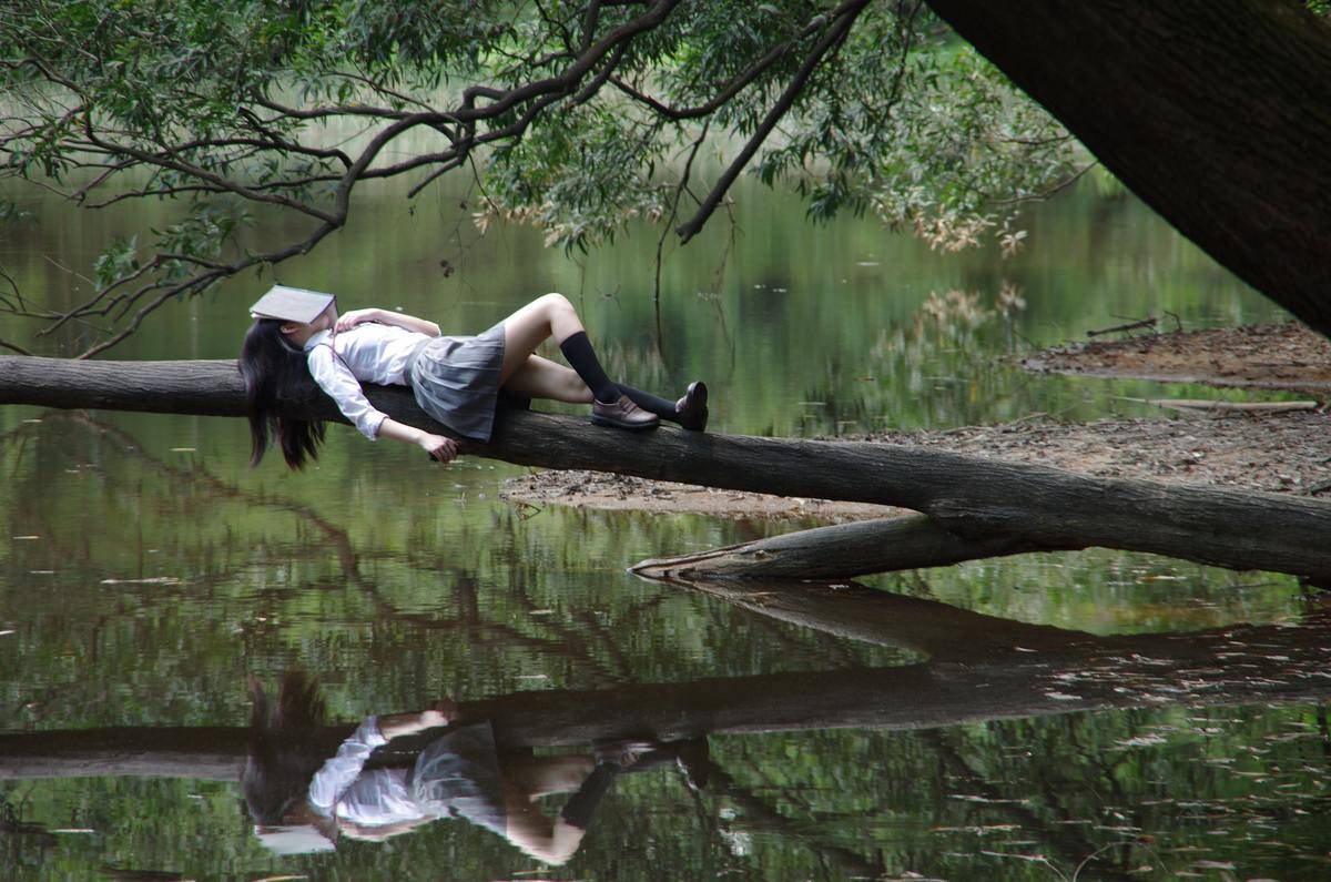 woman laying on branch with book on her face and reflection in the water
