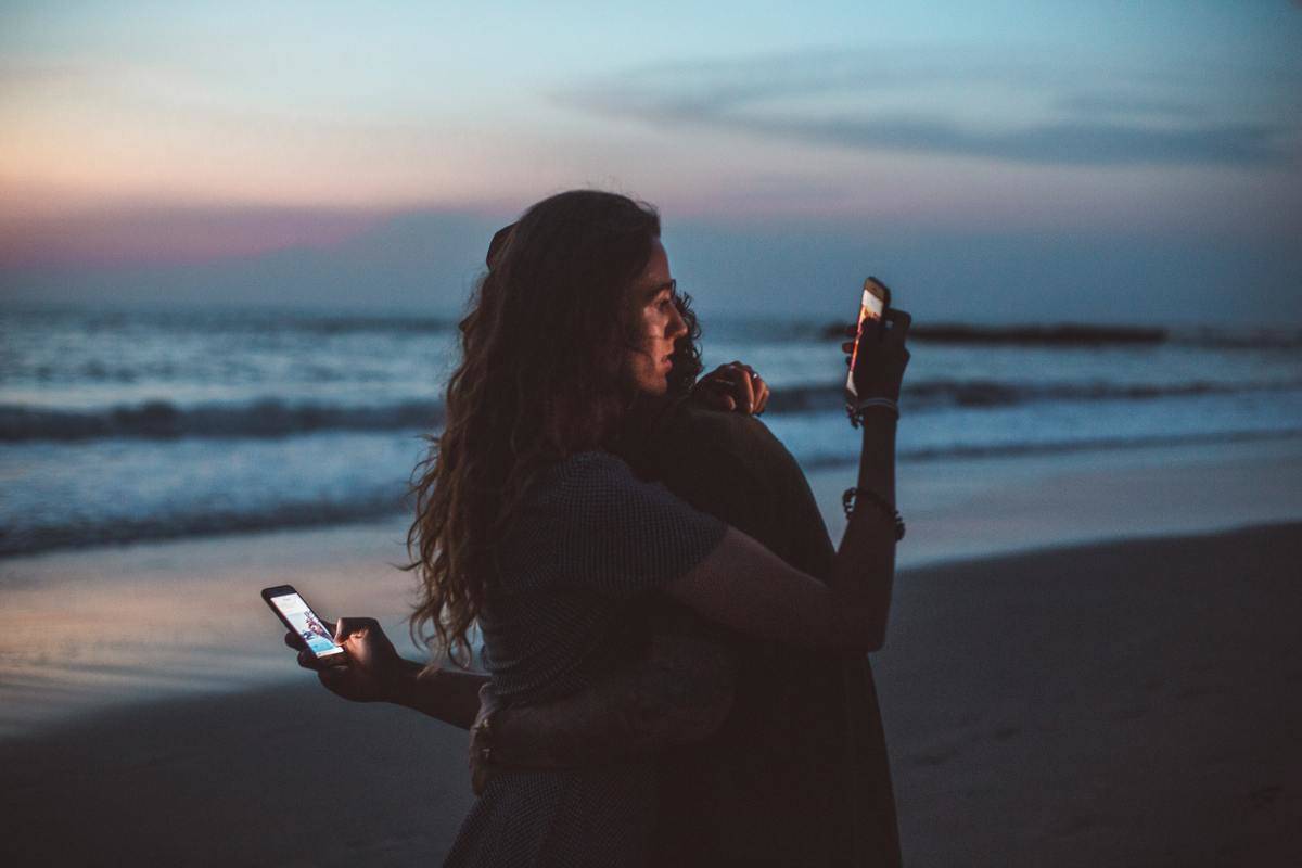 couple-hugging-and-using-smartphone-near-sea-on-sunset-facing away from each other