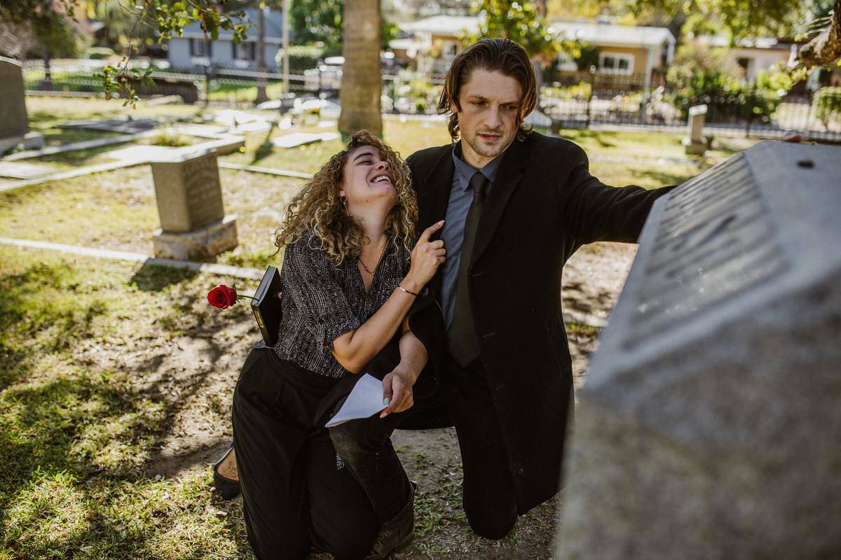 man-in-black-suit-jacket-sitting-beside-woman-in-black-and-white-shirt-and-black-pants- crying at cemetury