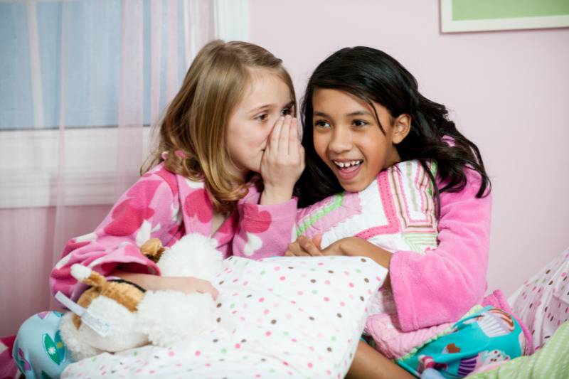 Young girl whisper to each other holding pillows in pjs on bed