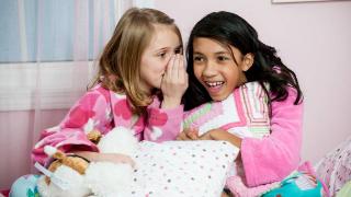 Young girl whisper to each other holding pillows in pjs on bed