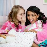 Young girl whisper to each other holding pillows in pjs on bed