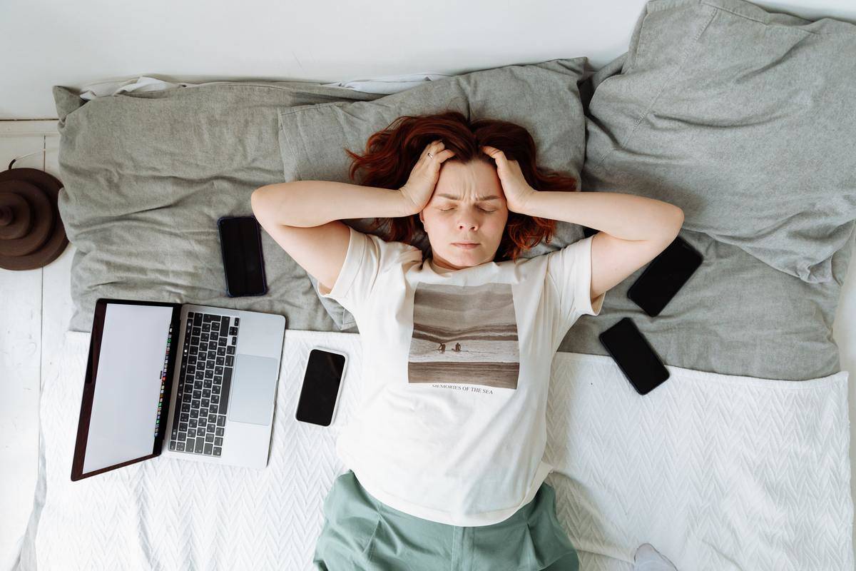 a-stressed-woman-lying-on-a-bed-beside-cellphones-and-a-laptop-