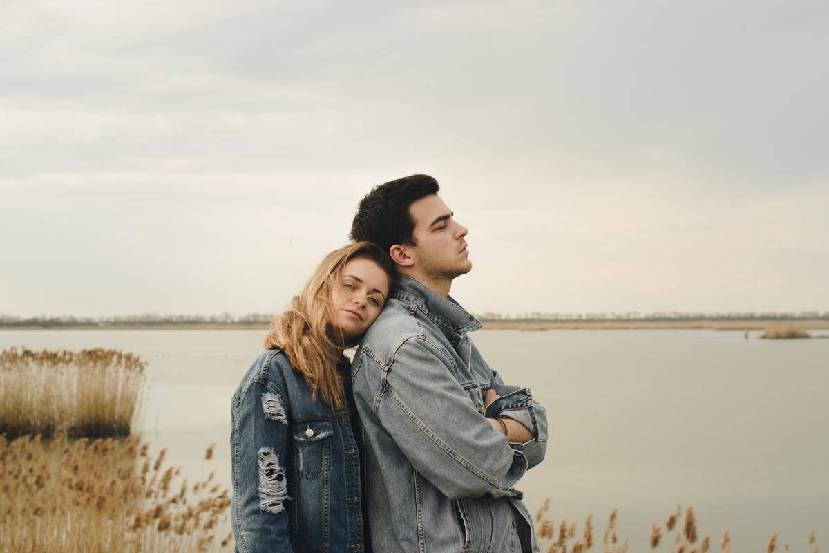 woman rests her head on man's shoulder who is facing away by pond, both in denim jackets