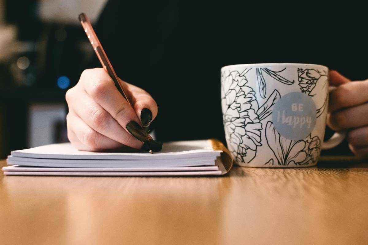 /person-holding-white-ceramci-be-happy-painted-mug-while writing