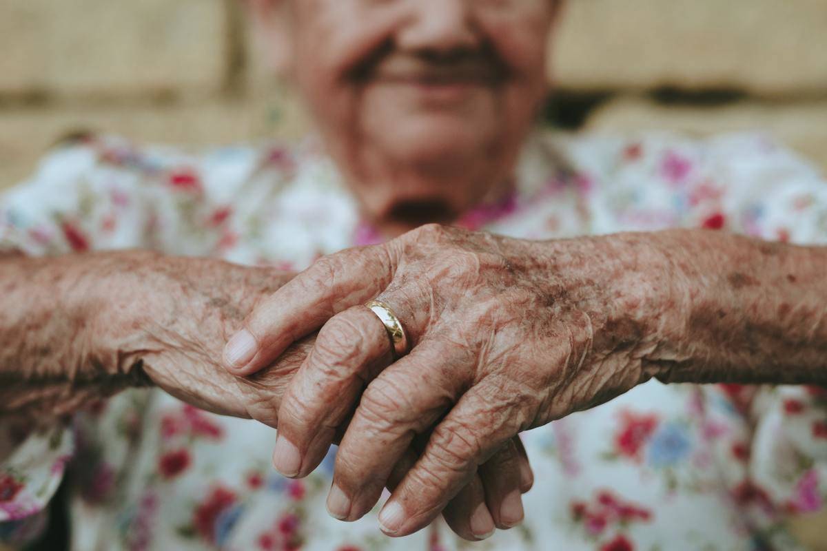 An elderly person with their hands folded together in the foreground.